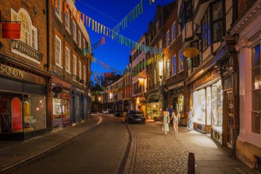 Cambridge, İngiltere - 12 Temmuz 2025: Cambridge 'de sıcak dükkan ışıkları, şenlik dekorları ve klasik bir eski kasaba ortamında romantik bir ortam olan kaldırımlı caddenin büyüleyici akşam manzarası.