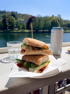 Sebzeli ve peynirli vejetaryen sandviç, güneşli bir günde bir bardak su ve bir gölün kenarındaki masada servis edilir..