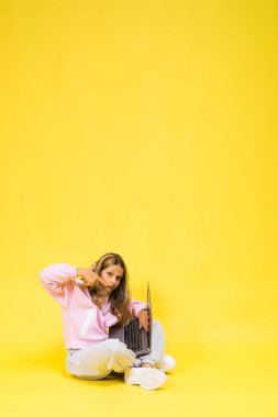 Pozitif çocuk elinde dizüstü bilgisayarla oturur, ödevini okur sarı arka planda günlük giysiler giyer.