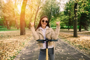 Sonbahar parkında elektrikli scooter kullanan genç bir kadın. Yeşil ulaşım, trafik sıkışıklığı.