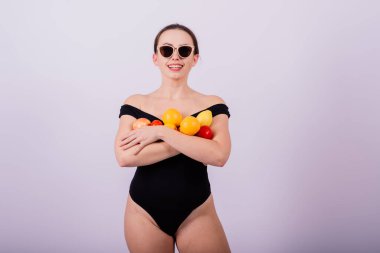 Siyah mayo giymiş, meyvelerle poz veren ve gülen güneş gözlüklü havalı bir kız..