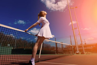 Sahada tenis raketi tutan beyaz elbiseli ve topuklu ayakkabılı seksi kız tenisçi. Genç kadın tenis oynuyor.