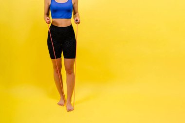 Formda bayan sporcu sarı arka plandaki fitness antrenmanında iple atlıyor.