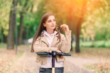 Sonbahar parkında elektrikli scooter kullanan genç bir kadın. Yeşil ulaşım, trafik sıkışıklığı.
