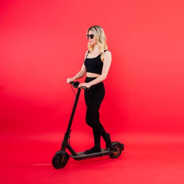 Kırmızı ve beyaz arka planda elektrikli scooterla spor kıyafetli güzel, duygusal genç bir bayan.