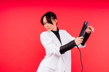 Gri ve kırmızı arka planda saç maşası tutan güzel saçlı mutlu genç kadın.