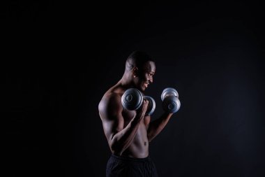 Isolated african muscular man with dumbbells on a dark studio background. Strong shirtless black guy