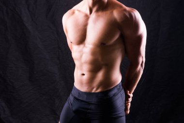 Çıplak göğsü olan bir adam karanlık bir zemine karşı poz veriyor, kaslı vücudunu gösteriyor. Flaşlı fotoğraflarda kafası, elleri, midesi, kolları, omuzları, bacakları ve boynu görünür.