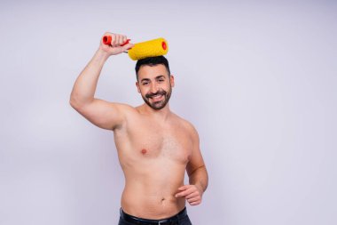 Neşeli genç üstsüz adam gri arka plan stüdyosunda izole edilmiş boya silindiri tutuyor..