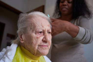 Afrikalı Amerikalı kadın bakıcı, evde yaşlı kadın saçını kesiyor.