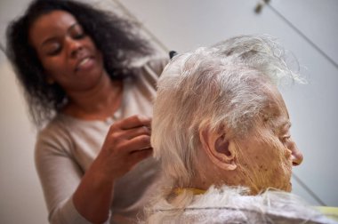 Afrikalı Amerikalı kadın bakıcı, evde yaşlı kadın saçını kesiyor.