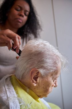 Afrikalı Amerikalı kadın bakıcı, evde yaşlı kadın saçını kesiyor.