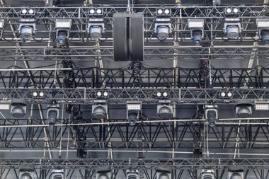 Birden fazla spot ışığı, LED ışıkları, hareket eden projektörleri ve hoparlörleri olan profesyonel bir sahne donanımının yakın çekimi. Fotoğraf, büyük ölçekli bir olayın arkasındaki teknik altyapıyı yakalıyor, hassasiyeti ve karmaşıklığı vurguluyor.