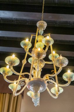 Rafine edilmiş cam avize, üzerinde altın detaylar var. Koyu ahşap kiriş tavandan sarkıyor. Parlayan ışıklar odayı hafifçe aydınlatarak sıcak ve zarif bir atmosfer yaratıyor. Gelenekleri birleştiren el işçiliği ve tasarımın mükemmel bir örneği. 