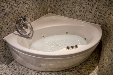 Modern otel banyosunda köpüklü suyla dolu bir jakuzi var. Küvet zarif mozaik fayanslarla çevrili, rafine ve rahatlatıcı bir spa atmosferi yaratıyor. Bu iç tasarım konforu, refahı ve lüksü ön plana çıkarıyor.