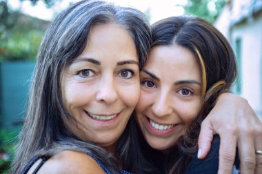 Gülümseyen bir anne ve yetişkin kızın yakın plan portresi. Sarılmak ve kameraya bakmak. Aile bağı, aşk ve mutluluk kavramı. Doğal ışık, açık hava..