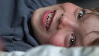 Yatakta gülümseyen 6 yaşındaki bir çocuğun yakın plan portresi. Yüksek kalite 4K görüntü. Mutlu şirin çocuk gülümseyip yatağında oynaşıyor. Yakından bir gülümseme, dişleri ve baklavaları..