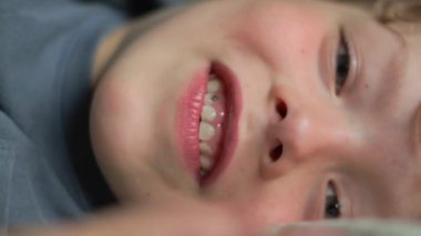 Yatakta gülümseyen 6 yaşındaki bir çocuğun yakın plan portresi. Yüksek kalite 4K görüntü. Mutlu şirin çocuk gülümseyip yatağında oynaşıyor. Yakından bir gülümseme, dişleri ve baklavaları..