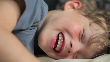 Yatakta gülümseyen 6 yaşındaki bir çocuğun yakın plan portresi. Yüksek kalite 4K görüntü. Mutlu şirin çocuk gülümseyip yatağında oynaşıyor. Yakından bir gülümseme, dişleri ve baklavaları..