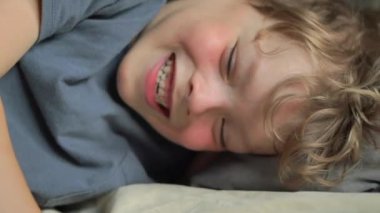 Yatakta gülümseyen 6 yaşındaki bir çocuğun yakın plan portresi. Yüksek kalite 4K görüntü. Mutlu şirin çocuk gülümseyip yatağında oynaşıyor. Yakından bir gülümseme, dişleri ve baklavaları..