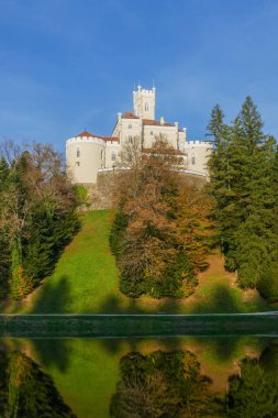 Trakoscan Kalesi, rengarenk sonbahar ormanlarıyla kaplı bir tepenin üzerine kurulmuş sakin bir gölün karşısından görülüyor. Resimli sahne, engin mavi gökyüzünün altındaki durgun suya yansıyor..
