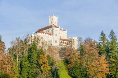 Kırmızı çatıları olan görkemli, krem renkli bir kale ve ormanlık bir tepenin üstünde merkezi bir kule bulunur. Çevreleyen ağaçlar açık mavi gökyüzünün altında sonbahar renklerini gösteriyor..