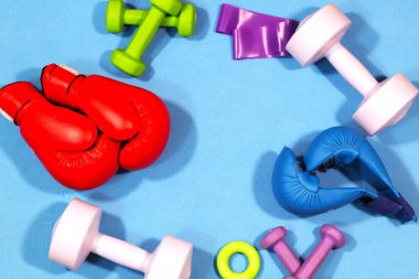 Parlak mavi bir paspasın üzerinde daire şeklinde dizilmiş düz renkli spor aletleri. Eşyalar arasında boks eldivenleri, dambıllar, direnç bantları ve fitness yüzükleri yer alıyor, dengeli ve dinamik bir yaşam tarzını sembolize ediyor.