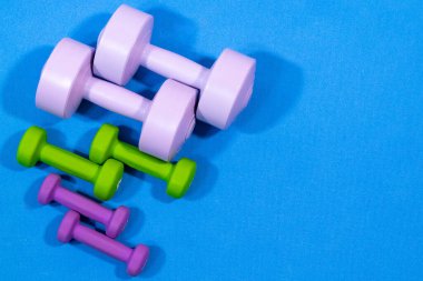Farklı boyutlarda renkli halterler seti (pembe, yeşil, mor) mavi bir egzersiz paspasına güzelce yerleştirilmiştir. Formun, spor salonunun ve sağlıklı yaşamın mükemmel bir temsilidir..