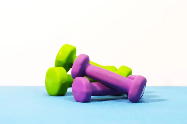 Yeşil ve mor halterlerin minimalist görüntüsü temiz beyaz arka planlı mavi egzersiz paspasına yerleştirilmiş. Fitness sadeliği, refah ve aktif yaşam temaları için mükemmel..