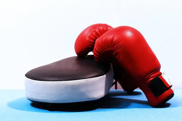 Parlak kırmızı boks eldivenleri siyah-beyaz bir yumruk yastığının üzerinde, temiz beyaz ve mavi bir arka planda duruyor. Görüntü, dövüş sanatları ve boksta hazırlık, eğitim ve sportmenlik içerir..