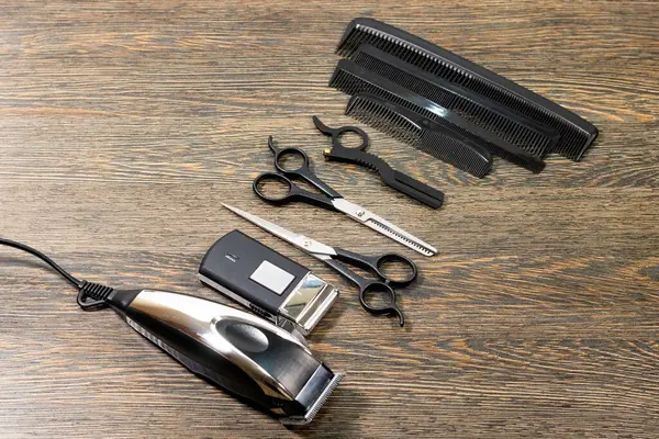 Bir elektrikli makas, kablosuz budama makinesi, çeşitli taraklar, iki çift makas ve seyreltici makas da dahil olmak üzere ahşap bir yüzeye düzgünce yerleştirilmiş profesyonel kuaför aletleri..