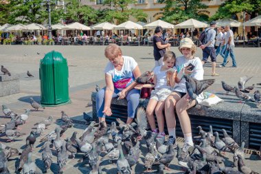Cracow, Poland: 23 July 2022: People feed pigeons, tourists attraction in Main Market Square in the Old city of Cracow, Poland.