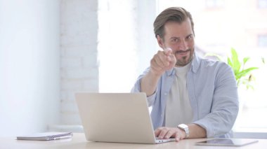 Mature Man Pointing at Camera while using Laptop