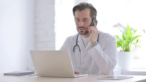Αρσενικό Γιατρός Μιλώντας Στο Τηλέφωνο Ενώ Εργάζονται Φορητό Υπολογιστή — Φωτογραφία Αρχείου