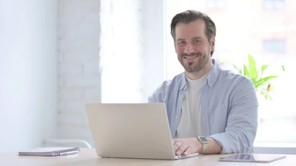 Ώριμος Άντρας Χαμογελάει Στην Κάμερα Ενώ Χρησιμοποιεί Laptop — Φωτογραφία Αρχείου