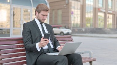 Middle Aged Businessman Using Smartphone and Laptop while Sitting Outdoor on Bench