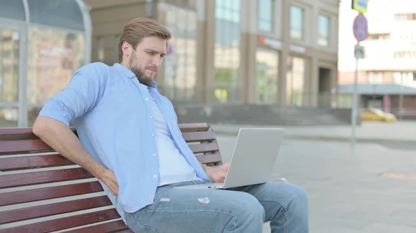 Middle Aged Man with Back Pain Using Laptop while Sitting Outdoor on Bench
