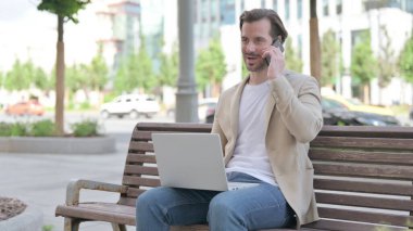 Man Talking on Phone and using Laptop while Sitting on Bench