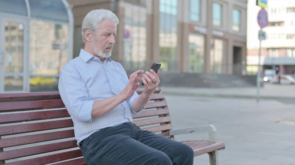 Senior Old Man Browsing Internet on Smartphone while Sitting Outdoor on Bench