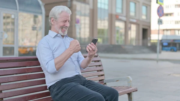 Senior Old Man Celebrating Online Success on Smartphone while Sitting Outdoor on Bench