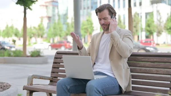 Angry Man with Laptop Talking on Phone while Sitting on Bench