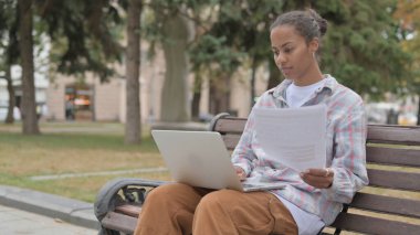 Genç Afrikalı Kadın Dokümanları okuyor ve Bench Outdoor 'da otururken dizüstü bilgisayarda çalışıyor.