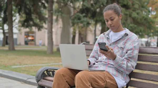 Young African Woman Using Smartphone and Laptop while Sitting Outdoor on Bench