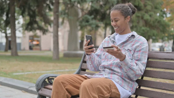 Young African Woman Shopping Online on Smartphone while Sitting Outdoor on Bench