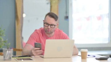 Genç Yetişkin Adam dizüstü bilgisayar kullanırken akıllı telefon kullanıyor