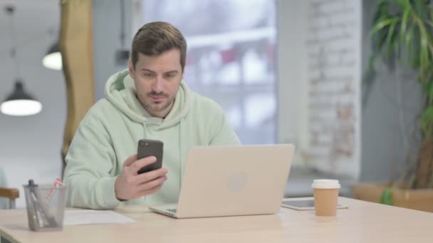 Genç Yetişkin Adam Dizüstü Bilgisayar Kullanırken Akıllı Telefon Kullanıyor — Stok video