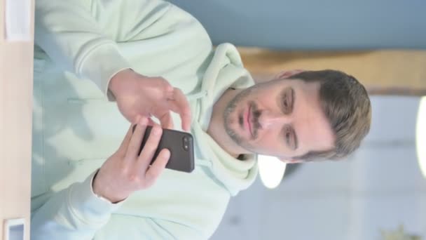 スマートフォンでインターネットを閲覧する若い大人の男の垂直ビデオ — ストック動画