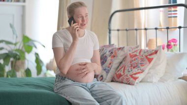 Pregnant Woman Talking on Phone while Sitting on Bed