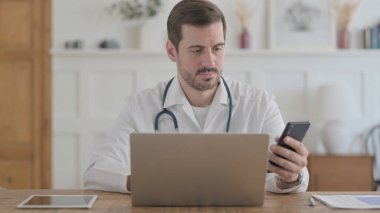 Male Doctor using Smartphone while using Laptop in Clinic