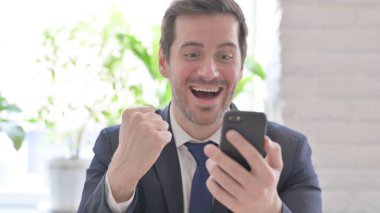 The Young Adult Businessman Celebrating Online Success on Smartphone in Office